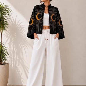 May include: A black open-front jacket featuring gold embroidered sun and moon motifs, worn over a white crop top and white wide-leg trousers with a drawstring waist. The text ZAHAAR is visible in the upper right corner of the image.