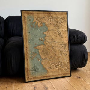 Könnte beinhalten: Ein gerahmtes, rechteckiges Wandbild mit einer detaillierten Fantasiekarte auf gealtertem, beigefarbenem Pergamentpapier. Die Karte zeigt blaue Küstengewässer, braune Gebirgszüge, Wälder und verschiedene beschriftete Orte mit einer Windrose.