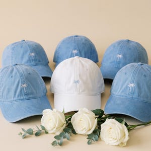 Könnte beinhalten: Eine Ansammlung von fünf Baseballkappen, die zusammen angeordnet sind. Vier Kappen sind hellblau mit weiß gestickten Palmenmotiven, und eine mittlere Kappe ist weiß mit einer hellblau gestickten Palme. Drei weiße Rosen liegen davor.
