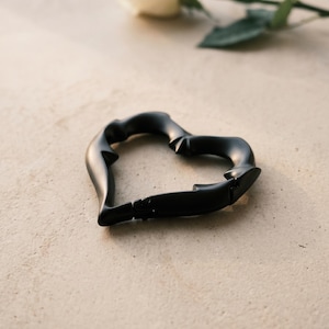 May include: A matte black heart-shaped carabiner with a smooth, contoured surface and a spring-loaded gate mechanism. The carabiner features a distinct, organic shape with subtle indentations along its frame, creating a sleek and functional accessory.
