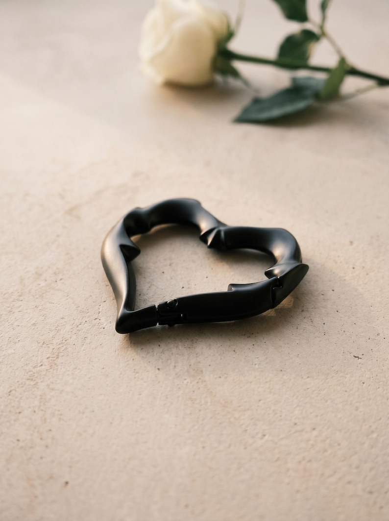 May include: A matte black heart-shaped carabiner with a smooth, contoured surface and a spring-loaded gate mechanism. The carabiner features a distinct, organic shape with subtle indentations along its frame, creating a sleek and functional accessory.