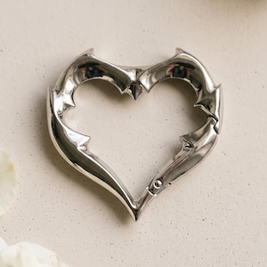 May include: A polished silver-tone metal carabiner shaped like a heart with sharp, pointed edges along the frame. The heart-shaped carabiner features a spring-loaded gate mechanism on the lower right side, allowing it to open and close securely.