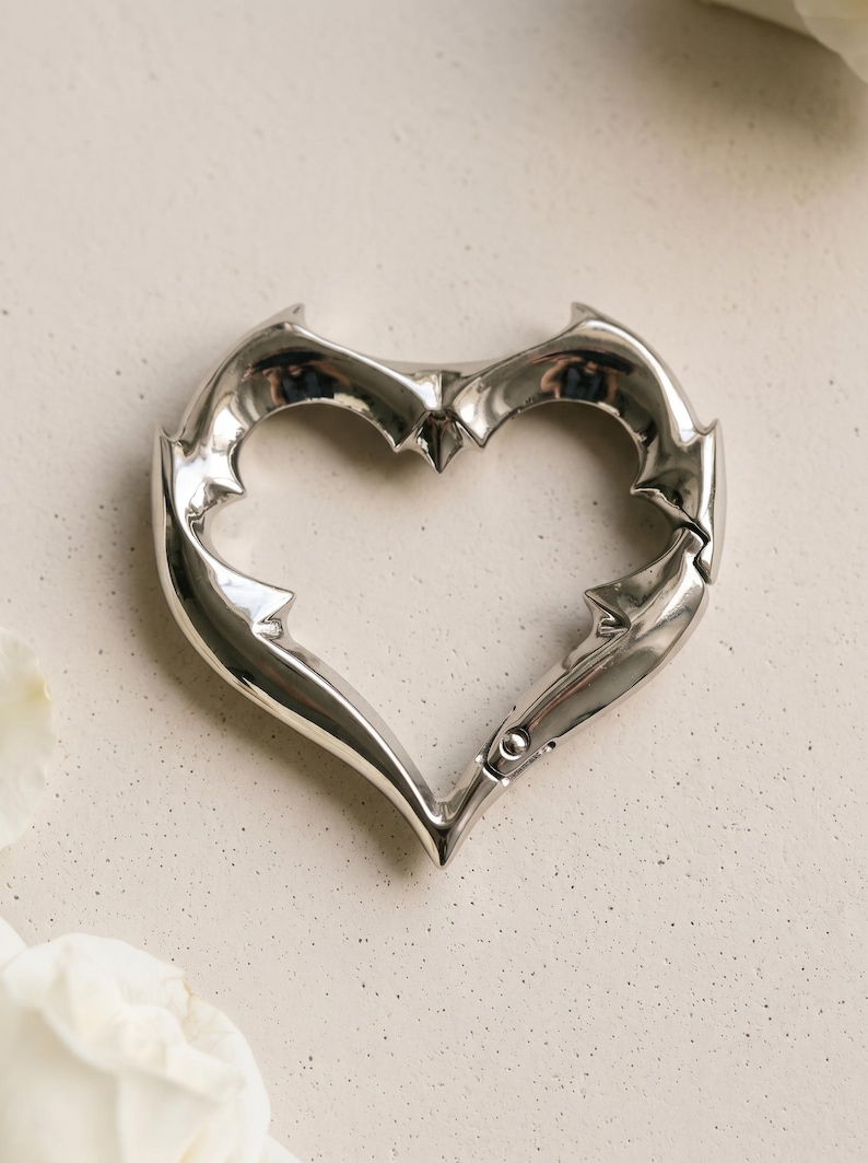 May include: A polished silver-tone metal carabiner shaped like a heart with sharp, pointed edges along the frame. The heart-shaped carabiner features a spring-loaded gate mechanism on the lower right side, allowing it to open and close securely.