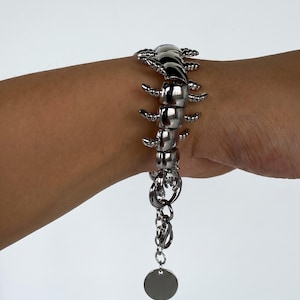 Könnte beinhalten: Ein silberfarbenes Metallarmband mit einem segmentierten, beweglichen Tausendfüßler-Design und mehreren Beinen an den Seiten. Das Armband verfügt über eine Verlängerungskette mit Karabinerverschluss und einen kleinen, runden, leeren Metallanhänger.
