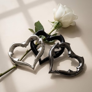 May include: Three heart-shaped carabiners with a thorny, jagged edge design. One carabiner is polished silver, one is matte black, and one is dark metallic grey. Each heart features a spring-loaded gate mechanism for opening and closing.