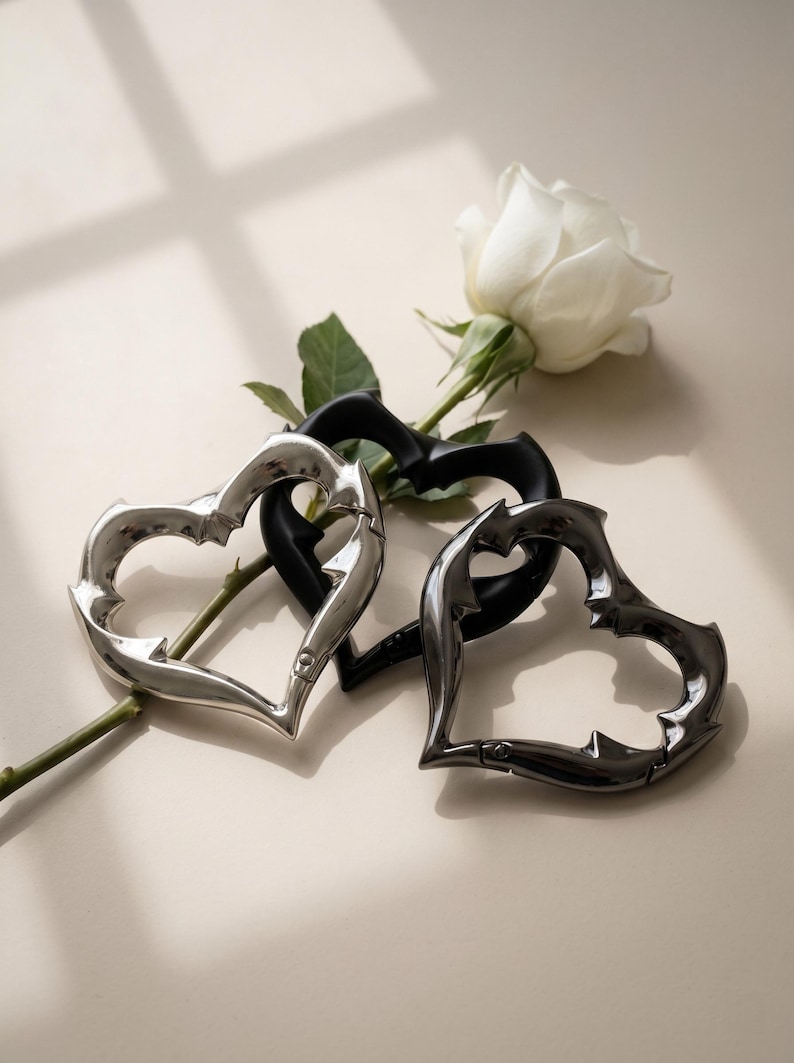 May include: Three heart-shaped carabiners with a thorny, jagged edge design. One carabiner is polished silver, one is matte black, and one is dark metallic grey. Each heart features a spring-loaded gate mechanism for opening and closing.