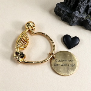 Könnte beinhalten: Ein goldfarbener Metallschlüsselanhänger mit einem menschlichen Skelettorso und Schädel an einem runden Ring. Ein runder, goldfarbener Metallanhänger hängt am Ring und ist mit dem Text Custom your Text With Logo graviert.