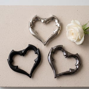 May include: Three heart-shaped carabiners with a thorny, spiked edge design. One carabiner is polished silver, one is matte black, and one is dark metallic grey. A single white rose with water droplets is positioned to the right of the hearts.