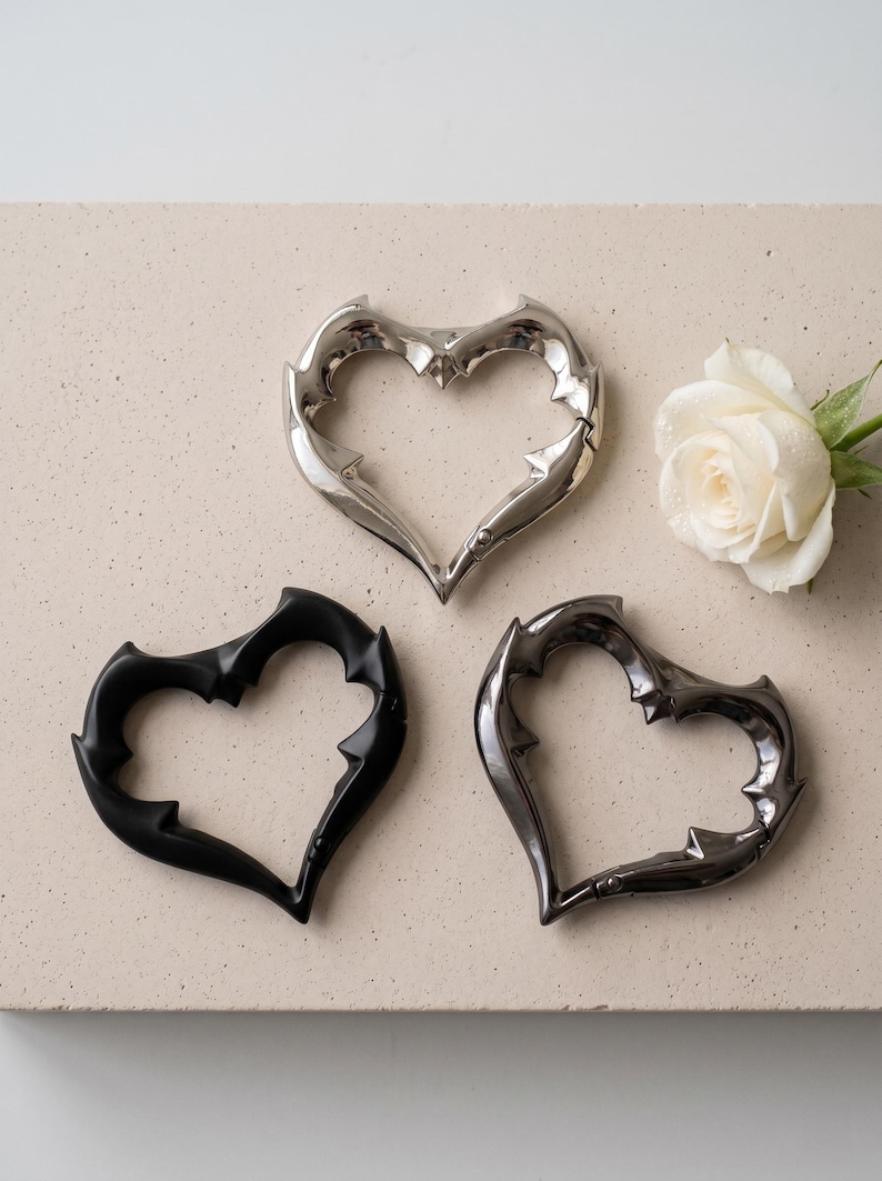 May include: Three heart-shaped carabiners with a thorny, spiked edge design. One carabiner is polished silver, one is matte black, and one is dark metallic grey. A single white rose with water droplets is positioned to the right of the hearts.