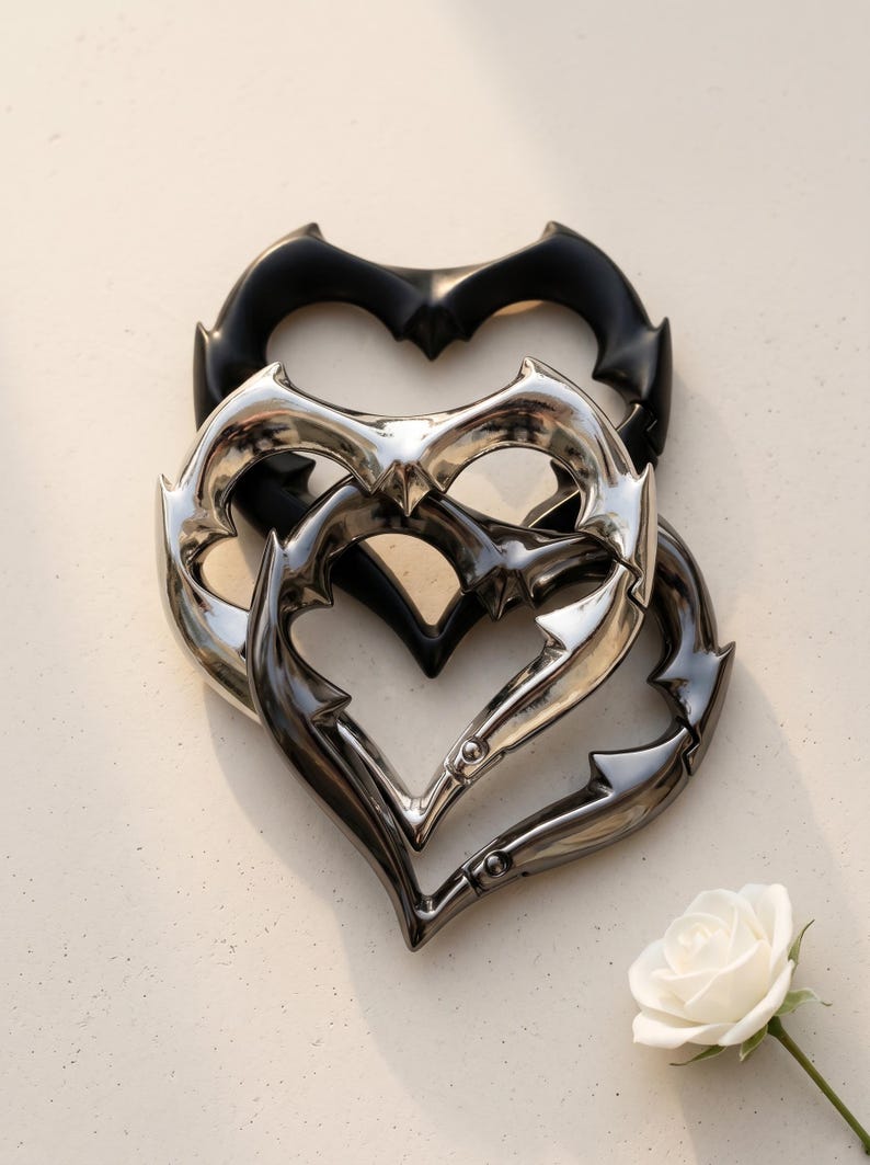 May include: Three heart-shaped carabiners with a spiked, thorny edge design. One carabiner is matte black, one is polished silver, and one is a dark gunmetal grey. Each features a spring-loaded gate mechanism for opening and closing the heart frame.