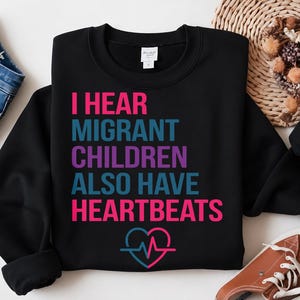 Peut inclure: Un sweat-shirt noir à manches longues avec le texte I HEAR MIGRANT CHILDREN ALSO HAVE HEARTBEATS en lettres roses, bleu sarcelle et violettes, accompagné d'un graphique en forme de cœur et de battement de cœur en rose et bleu sarcelle.