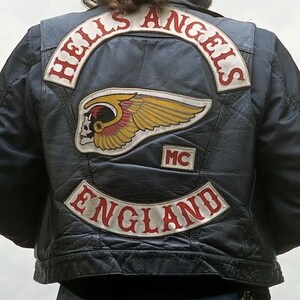 Może przedstawiać: Czarna skórzana kamizelka z dużą naszywką na plecach z napisem HELLS ANGELS w kolorze czerwonym na białym tle, centralnym emblematem uskrzydlonej czaszki w kolorze żółtym i czerwonym, małą naszywką MC oraz dolną naszywką z napisem ENGLAND w kolorze czerwonym.