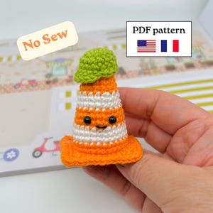 Puede incluir: Un pequeño cono de tráfico de ganchillo con rayas horizontales naranjas y blancas, una gorra verde en la parte superior y dos ojos de cuentas negras. El texto dice No Sew y PDF pattern junto a banderas de Estados Unidos y Francia.