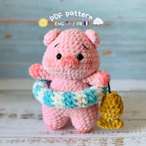 Peut inclure: Une peluche cochon crochetée rose avec des yeux noirs, portant une bouée en fil bleu et blanc autour de la taille et tenant un petit poisson crocheté jaune. Le texte indique PDF pattern ENG / FR.