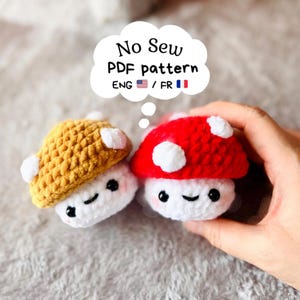 Peut inclure: Deux petits champignons en crochet avec des pieds blancs et des chapeaux jaune moutarde et rouge vif, ornés de taches blanches, d'yeux en perles noires et de sourires brodés. Une bulle indique : No Sew PDF pattern ENG 🇺🇸 / FR 🇫🇷.