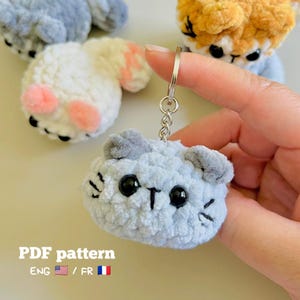 Peut inclure: Un porte-clés chat crocheté bleu clair avec des yeux en plastique noir et des moustaches brodées, muni d'un anneau métallique. Le texte PDF pattern ENG est visible en bas à gauche, avec un drapeau américain et un drapeau français.