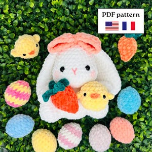 Peut inclure: Un lapin au crochet blanc avec des oreilles pêche tenant une carotte orange, accompagné d'un poussin jaune et de plusieurs œufs au crochet colorés. Un panneau blanc avec le texte PDF pattern et les drapeaux américain et français est visible.