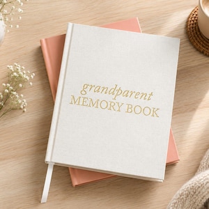May include: A white linen-textured hardback book with the gold-stamped text "grandparent MEMORY BOOK" on the front cover, featuring a white ribbon bookmark, resting on top of a solid peach-coloured book.