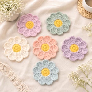 May include: Six round crochet coasters shaped like flowers, each featuring a bright yellow centre surrounded by scalloped petals in pastel shades of light pink, mint green, cream, peach, lavender, and light blue, arranged on a fabric surface.