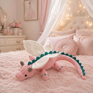 May include: A handmade crochet dragon plush toy featuring a soft pink body, large white wings, and a dark teal spiked ridge running along its back and tail. The creature has small grey horns and grey accents on its feet, with black bead eyes.