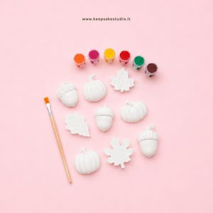 Könnte beinhalten: Ein Bastelset mit weißen Gipsfiguren in Form von Kürbissen, Eicheln und Blättern, dazu ein Holzpinsel und sieben kleine runde Farbtöpfe in Orange, Pink, Gelb, Rot, Grün und Braun. Der Text www.keepsakestudio.it ist auf dem Bild zu sehen.