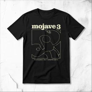 Puede incluir: Una camiseta negra de manga corta con un gráfico de líneas color crema de una figura y una guitarra dentro de un marco cuadrado. El texto "mojave 3" aparece sobre el marco y el texto "excuses for travellers" está impreso debajo del marco.