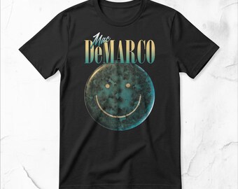 Op de afbeelding: Een zwart t-shirt met korte mouwen en de tekst Mac DeMARCO in een blauw en geel verlooplettertype boven een cirkelvormige afbeelding. De afbeelding toont een getextureerd, donkerblauwgroen smileygezicht met twee kleine gele stippen en een boog.
