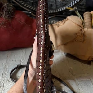 May include: A slim, dark brown leather crossbody bag featuring a woven pattern with visible light-coloured stitching along the edges. The bag has a narrow profile, a thin adjustable shoulder strap, and a metallic clasp closure on the side.