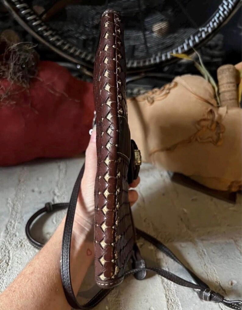 May include: A slim, dark brown leather crossbody bag featuring a woven pattern with visible light-coloured stitching along the edges. The bag has a narrow profile, a thin adjustable shoulder strap, and a metallic clasp closure on the side.