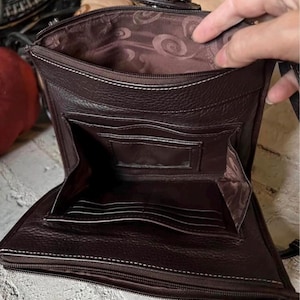 May include: A dark brown leather crossbody bag with a zippered main compartment and a front flap pocket. The interior features a brown fabric lining with a swirling pattern, multiple card slots, and a clear identification window pocket.