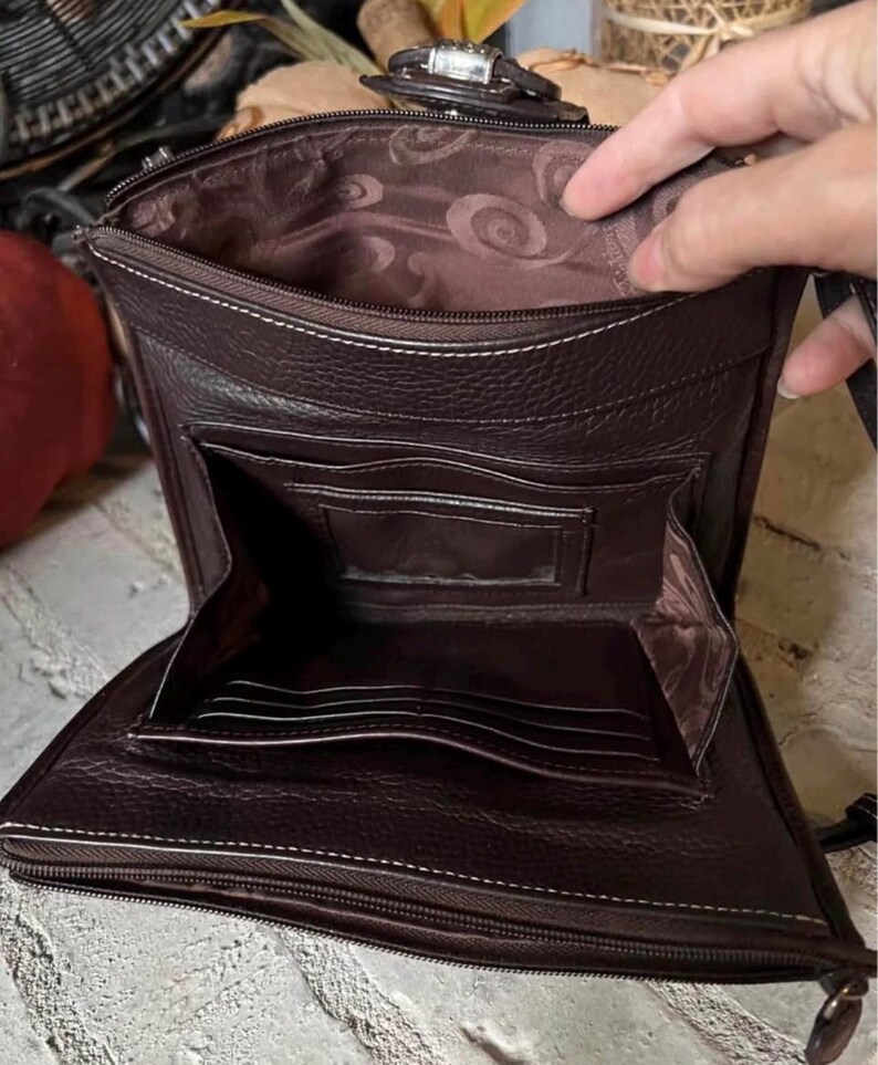 May include: A dark brown leather crossbody bag with a zippered main compartment and a front flap pocket. The interior features a brown fabric lining with a swirling pattern, multiple card slots, and a clear identification window pocket.