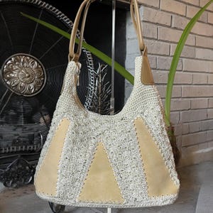 May include: A hobo-style shoulder bag featuring a cream-colored crocheted body with a textured shell pattern. The bag is accented with three vertical, smooth tan leather panels and includes matching tan leather shoulder straps and trim details.