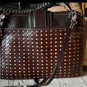 May include: A dark brown rectangular crossbody bag featuring a textured leather upper section and a lower panel with a laser-cut heart pattern revealing a light beige fabric lining underneath. The bag includes a top zipper and a thin shoulder strap.