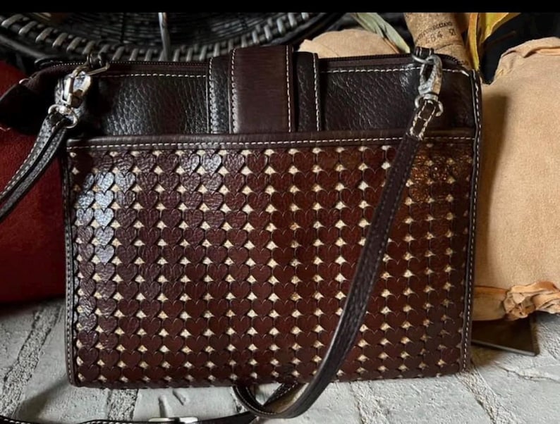 May include: A dark brown rectangular crossbody bag featuring a textured leather upper section and a lower panel with a laser-cut heart pattern revealing a light beige fabric lining underneath. The bag includes a top zipper and a thin shoulder strap.