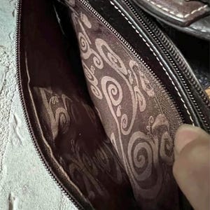 May include: The interior of a dark brown bag featuring a dark brown fabric lining with a repeating light brown swirl pattern. The bag has a zipped closure with a metal zip track and a dark brown leather-like exterior trim with white stitching.