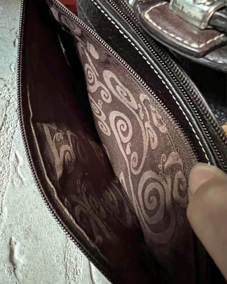 May include: The interior of a dark brown bag featuring a dark brown fabric lining with a repeating light brown swirl pattern. The bag has a zipped closure with a metal zip track and a dark brown leather-like exterior trim with white stitching.
