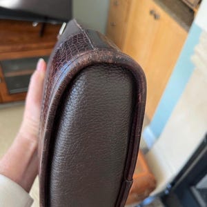 以下が含まれることがあります： 本体に質感のある粒状の表面を持ち、丸みを帯びたエッジに沿ってコントラストのあるダークブラウンのクロコダイル型押しレザーのトリムが施されたダークブラウンのレザーバッグ。レザーのパイピング部分には目に見える摩耗や擦り傷がある。