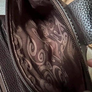 May include: A dark brown handbag with a textured exterior and a zip closure. The interior is lined with a brown fabric featuring a repeating swirling pattern. The bag includes a dark brown strap attached to the side with a metallic ring hardware.