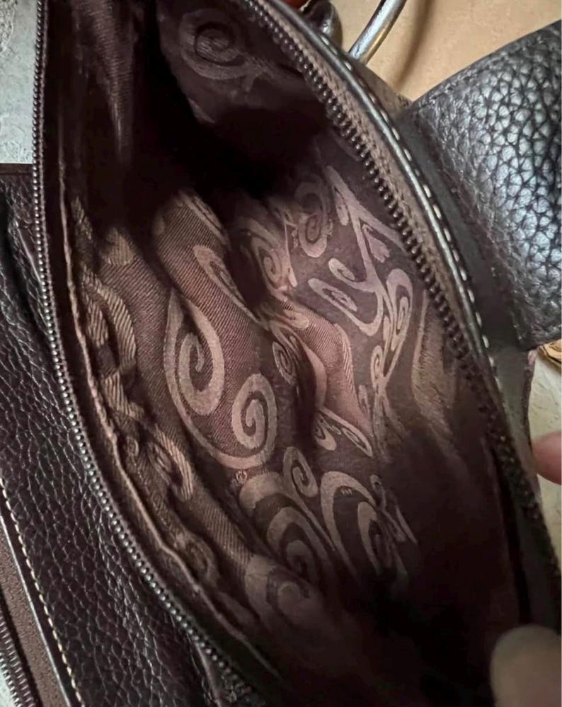 May include: A dark brown handbag with a textured exterior and a zip closure. The interior is lined with a brown fabric featuring a repeating swirling pattern. The bag includes a dark brown strap attached to the side with a metallic ring hardware.