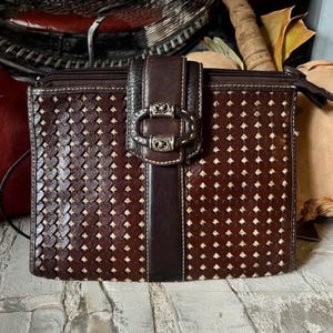 May include: A rectangular dark brown handbag featuring a repeating heart-shaped cutout pattern over a lighter tan base. The bag includes a central vertical strap with a decorative silver-toned metal buckle closure and a top zipper opening.