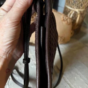May include: A dark brown crossbody bag featuring a woven leather texture on the side panel, a slim profile, and a thin adjustable shoulder strap with silver-toned metal hardware. The bag has visible stitching along the edges and a smooth leather finish.