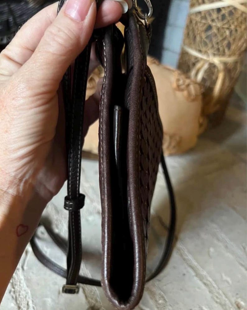 May include: A dark brown crossbody bag featuring a woven leather texture on the side panel, a slim profile, and a thin adjustable shoulder strap with silver-toned metal hardware. The bag has visible stitching along the edges and a smooth leather finish.