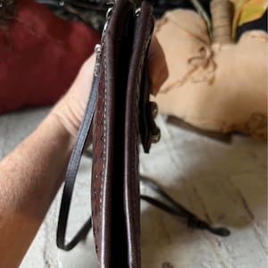May include: A slim, dark brown leather crossbody bag with a textured, embossed pattern on the side panel. The bag features a thin, matching leather shoulder strap and a visible metal hardware attachment point on the side edge of the structure.