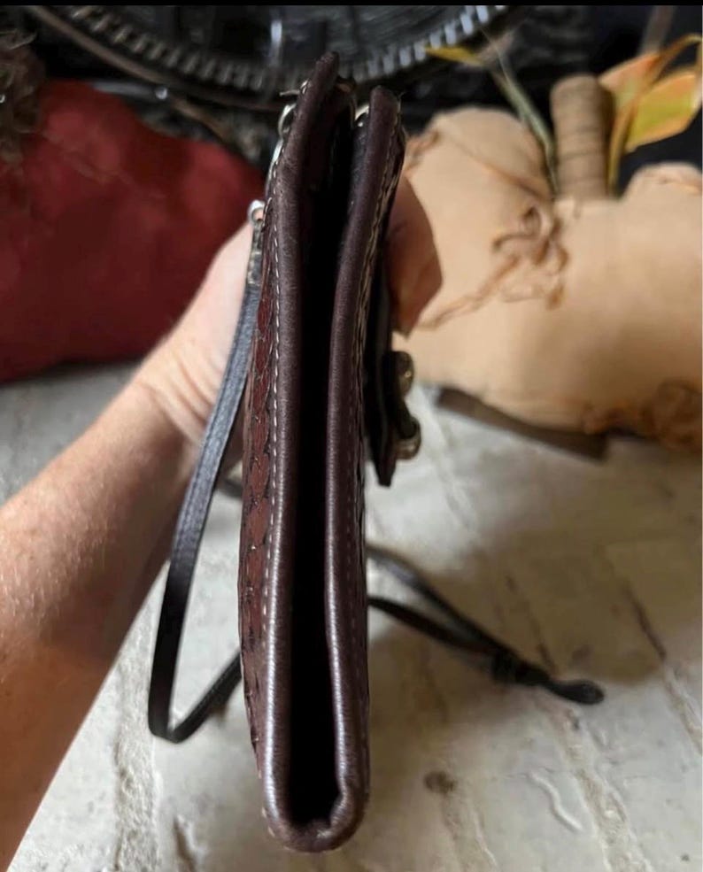 May include: A slim, dark brown leather crossbody bag with a textured, embossed pattern on the side panel. The bag features a thin, matching leather shoulder strap and a visible metal hardware attachment point on the side edge of the structure.