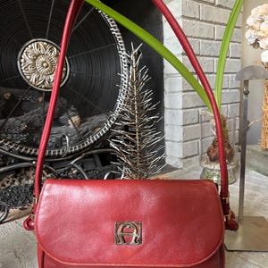 May include: A red leather shoulder bag featuring a single long strap with a knotted top detail and a gold-toned metal emblem on the front flap. The bag has a rectangular shape with rounded corners and gold-toned hardware connecting the strap.