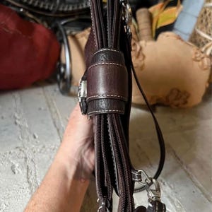 May include: A dark brown leather crossbody bag featuring multiple zippered compartments, a central magnetic snap closure flap with visible white stitching, and silver-toned metal hardware attachments for a shoulder strap.