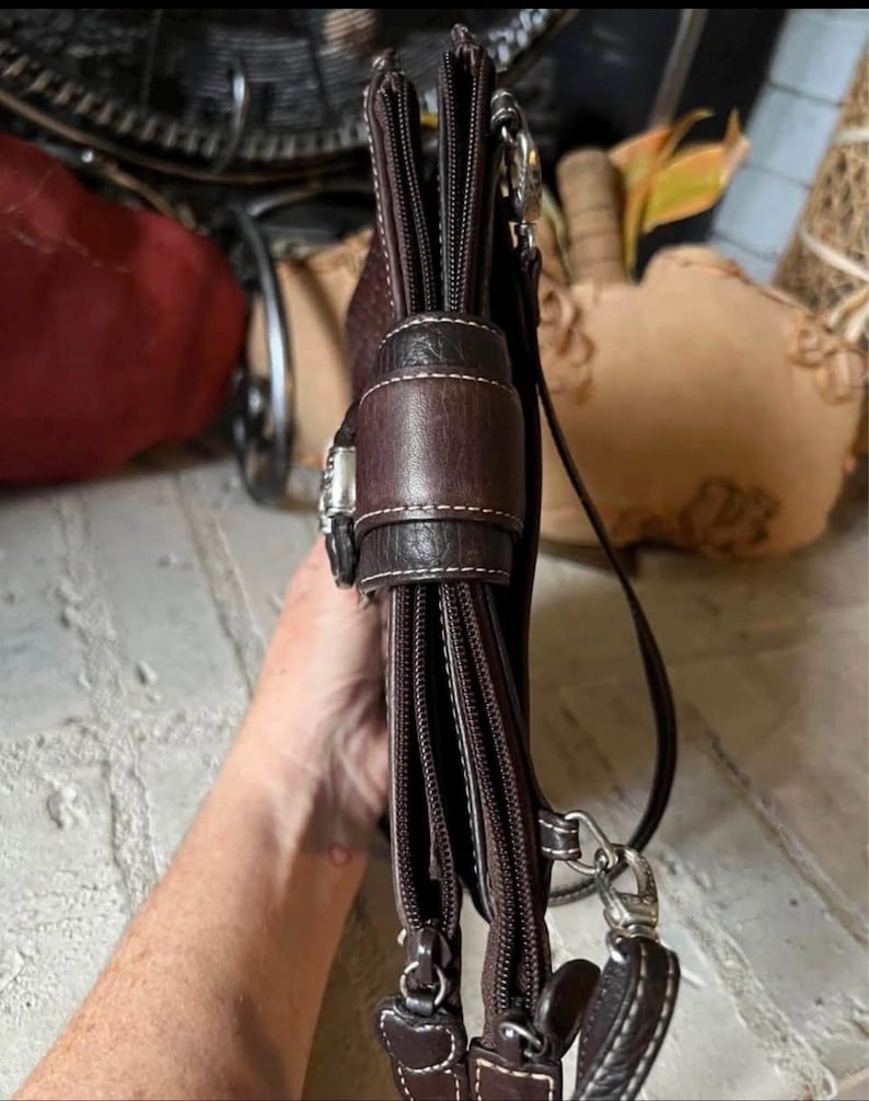 May include: A dark brown leather crossbody bag featuring multiple zippered compartments, a central magnetic snap closure flap with visible white stitching, and silver-toned metal hardware attachments for a shoulder strap.