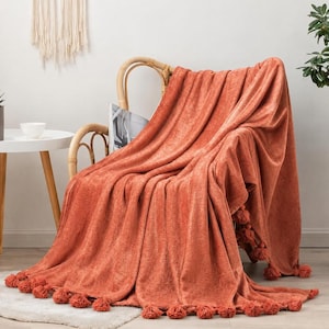 May include: A solid burnt orange throw blanket draped over a light-coloured wooden chair. The blanket features a soft, textured fabric and is finished with a decorative row of matching orange tassels along the bottom edge, creating a draped effect.