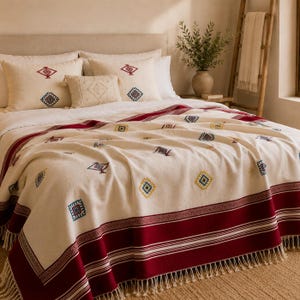 May include: An off-white bedspread featuring a deep red border with thin white stripes and fringe trim. The fabric is decorated with small, embroidered geometric diamond patterns in red, blue, and yellow, matching three decorative throw pillows.