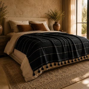 May include: A dark charcoal throw blanket with a subtle white grid pattern and black tassels along the bottom edge, draped over a bed with beige bedding, two brown accent pillows, and two cream-colored pillows in a warm, sunlit bedroom setting.